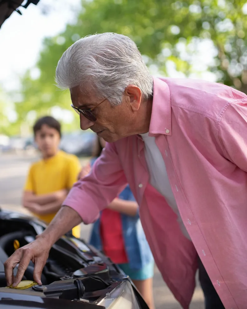 Idősebb, rózsaszín inges férfi hajol egy autó nyitott motortere fölé egy fás utcán, a háttérben gyerekek
